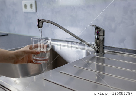 Water is running from the tap in the kitchen. A girl takes water into a glass from the tap in the kitchen 120096787