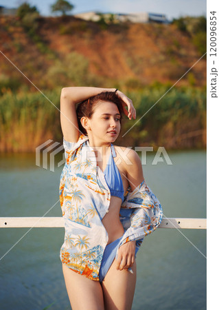 Cute slim young woman in blue swimsuit and Hawaiian shirt on a bridge by river 120096854