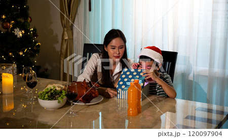 Mother and son enjoying Christmas together, with boy in Santa hat opening gift box at decorated holiday dinner table 120097904