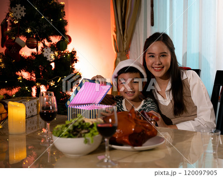 Happy mother and son celebrating Christmas, sitting at festive dinner table with glowing gift box and Christmas tree Happy mother and son celebrating Christmas, sitting at festive dinner table with glowing gift box and Christmas tree 120097905