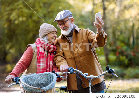 Lovely elderly couple on standing in the middle of autumn nature with their bikes, taking selfie with smartphone. Lovely elderly couple on standing in the middle of autumn nature with their bikes, taking selfie with smartphone. 120099109