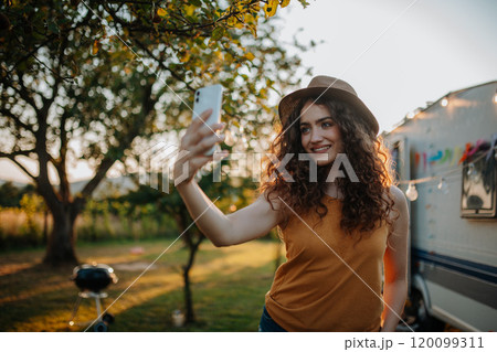 Portrait of young beautiful woman on a camping trip in nature, standing in front of caravan with smartphone in hand, taking selfie photo. 120099311