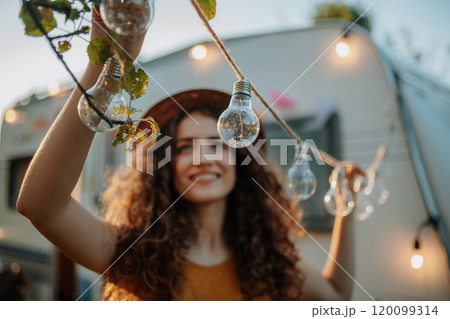 Young beautiful woman is on a camping trip in nature, putting lights on tree, preparing for evening. Young beautiful woman is on a camping trip in nature, putting lights on tree, preparing for evening. 120099314