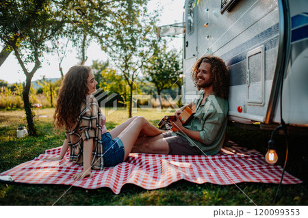 Couple sitting by caravan, enjoying peaceful moment. Man playing guitar and singing song to his girlfriend. Camping trip for young people. 120099353