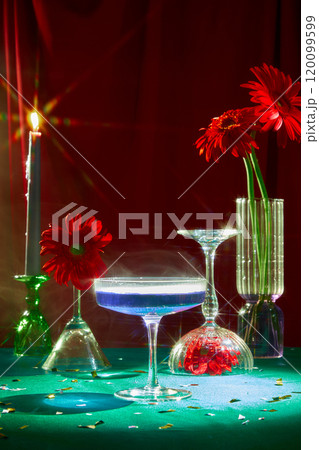 Party-themed still life shot, close-ups of cocktail glasses, a candle holder with a light candle, a glass vase, and some red flowers on a green surface against a red backdrop. 120099599