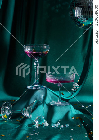 A party theme template with a front view of a raised green surface displays cocktail glasses filled with purple water, a glass resting next to ice, and confetti. From above, a disco ball is suspended. 120099600