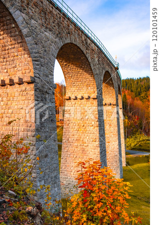 On a sunny autumn day, the railway viaduct in Smrzovka showcases stunning stone arches surrounded by vibrant fall foliage, creating a picturesque landscape in the Jizera Mountains of Czechia. 120101459