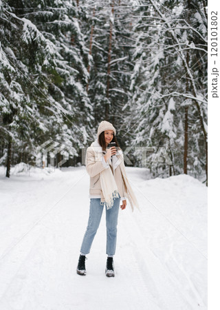 Happy woman is walking in snow with scarf wrapped around her neck and hat on her head. She is holding up of coffee in her hand 120101802