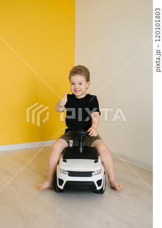 Boy child sits in toy car and gives thumbs up. Scene is bright and cheerful, with yellow wall and smile 120101803