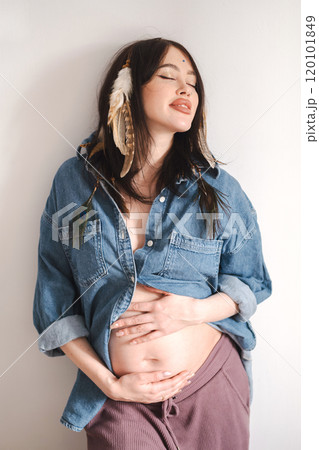 Pregnant woman poses in denim shirt with feathers in her hair, celebrating motherhood. Pregnant woman poses in denim shirt with feathers in her hair, celebrating motherhood. 120101849