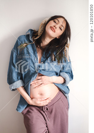 Pregnant woman poses in denim shirt with feathers in her hair, celebrating motherhood. Pregnant woman poses in denim shirt with feathers in her hair, celebrating motherhood. 120101850