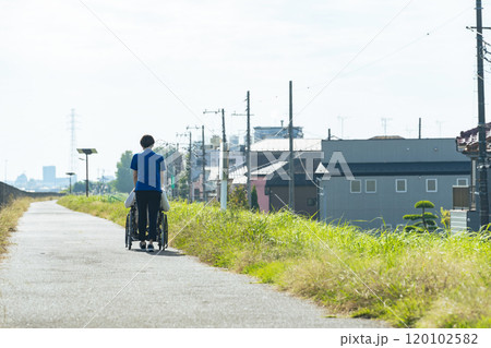 街を歩く車椅子に乗ったシニア男性と女性介護士の後ろ姿 120102582