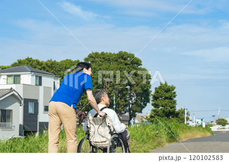 街を歩く車椅子に乗ったシニア男性と介護士の後ろ姿 120102583