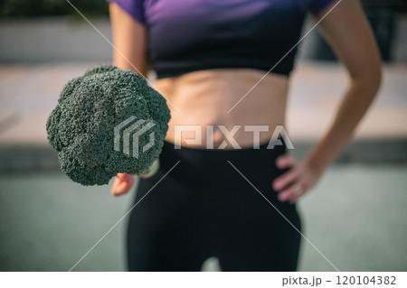 A woman holds a large piece of broccoli while posing confidently in athletic wear outdoors A woman holds a large piece of broccoli while posing confidently in athletic wear outdoors 120104382