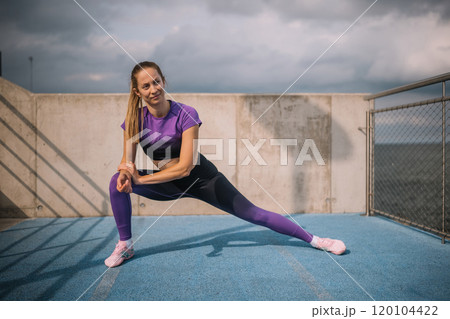 A fit woman stretches outdoors on a blue track near the ocean under cloudy skies in the afternoon A fit woman stretches outdoors on a blue track near the ocean under cloudy skies in the afternoon 120104422