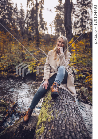 A woman in coat and jeans relaxes on a fallen tree by a serene stream in autumn A woman in coat and jeans relaxes on a fallen tree by a serene stream in autumn 120104493