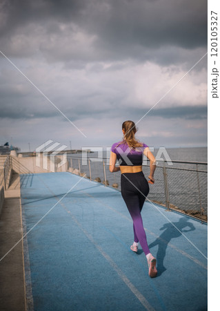 Woman jogging along a coastal path on a cloudy day, with a blue running track and ocean view 120105327