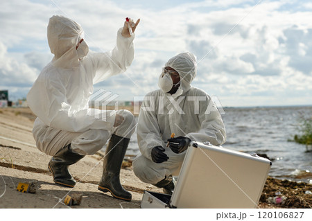 Full length side view of two scientists taking probes outdoors in nature and holding test tubes while wearing protective suits by water, copy space 120106827