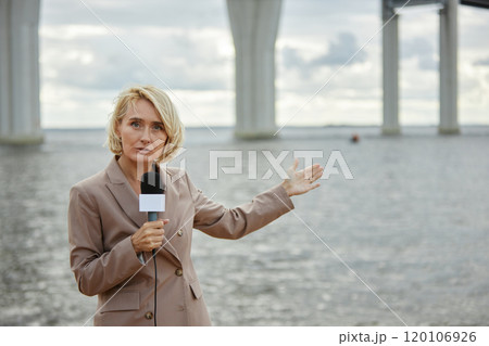 Waist up portrait of young blonde woman as news reporter speaking to microphone and gesturing to vast water landscape in background, copy space 120106926