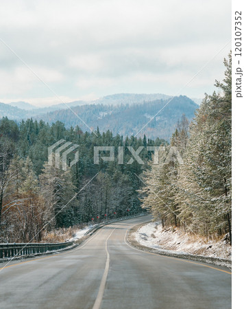 Winter road in snowy mountains. Autumn or wintertime in mountains. Asphalted road through fir tree forest and white snowy mountain peaks in the distance on background. Amazing landscape. Copy space 120107352
