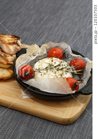 Grilled Camembert cheese with tomatoes and toasted bread. Camembert in a grill pan on a wooden board. Grilled Camembert cheese with tomatoes and toasted bread. Camembert in a grill pan on a wooden board. 120107503