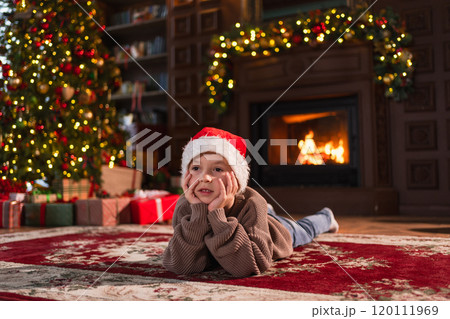 Merry Christmas. Little boy smiling near Christmas tree in classical dark interior. Young happy kid wearing Santa Hat in living room with fireplace Christmas tree gift boxes. Christmas eve at home 120111969