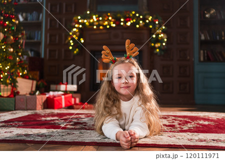 Merry Christmas. Little girl smiling near Christmas tree in classical dark interior. Young happy kid wearing deer horns in living room with fireplace Christmas tree gift boxes. Christmas eve at home 120111971