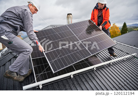 Builders installing solar panel system on roof of house. Men workers in helmets carrying photovoltaic solar module outdoors. Concept of alternative and renewable energy. Builders installing solar panel system on roof of house. Men workers in helmets carrying photovoltaic solar module outdoors. Concept of alternative and renewable energy. 120112194