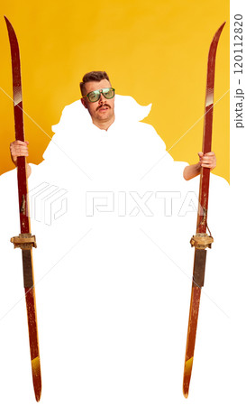 Man wearing vintage ski goggles and holding old-fashioned wooden skis against bright yellow background with blank white space. 120112820