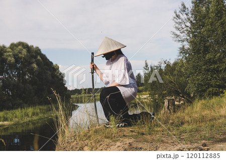 A female samurai in traditional attire kneels by the river holding a katana. The tranquil landscape encourages reflection on Japanese culture as the samurai warrior holds a katana. 120112885