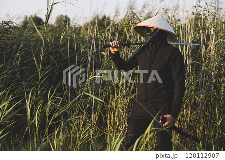 A samurai wearing a mask, conical hat and black clothes holds a katana over his shoulder while standing in a bamboo forest, possibly practicing swordsmanship. Samurai with katana. 120112907