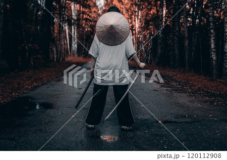 A samurai warrior wearing a straw hat stands on a path in the forest holding a katana. The warrior with the katana is illuminated by the sun setting in the background. 120112908