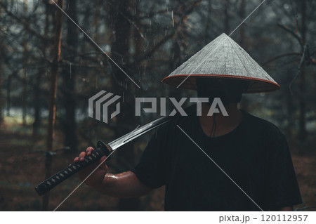 A samurai with a katana stands in the rain in the forest. A mysterious swordsman in a conical Asian hat and a mask on his face, holding a katana on his shoulder, stands in a dark rainy forest. A samurai with a katana stands in the rain in the forest. A mysterious swordsman in a conical Asian hat and a mask on his face, holding a katana on his shoulder, stands in a dark rainy forest. 120112957