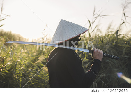 A samurai in traditional clothing carries a katana at sunset in a field bathed in golden light, evoking the spirit of the Japanese warrior. The samurai stands with the katana slung over his shoulder. A samurai in traditional clothing carries a katana at sunset in a field bathed in golden light, evoking the spirit of the Japanese warrior. The samurai stands with the katana slung over his shoulder. 120112973