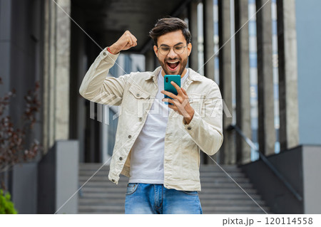 Indian man use mobile smartphone celebrating win good message news outdoors in urban city street Indian man use mobile smartphone celebrating win good message news outdoors in urban city street 120114558