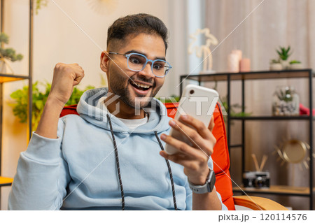 Indian Arabian man at home use smartphone celebrating success victory winning play online games 120114576