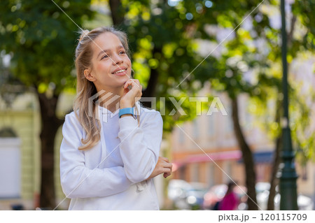 Thoughtful smiling young woman with pensive expression touching chin looking around on city street 120115709