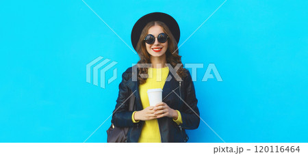 Stylish happy young woman holding cup of coffee in black hat, leather jacket on blue background 120116464