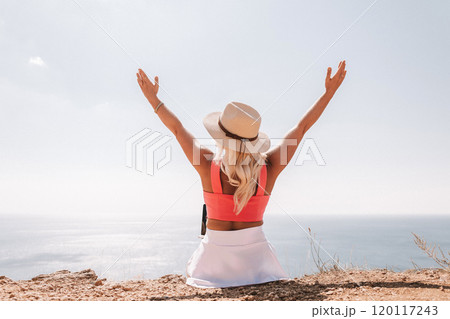 A woman wearing a pink top and a straw hat is sitting on a beach, looking out at the ocean. She is smiling and she is enjoying the view. 120117243