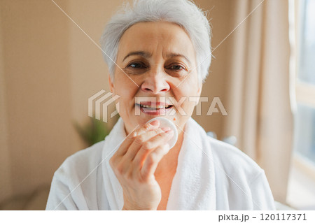 Mature adult old woman doing daily beauty routine removing makeup with cotton pad. Senior woman with lotion sponge washing face gets facial treatment. Skin care aging beauty skincare cleansing 120117371