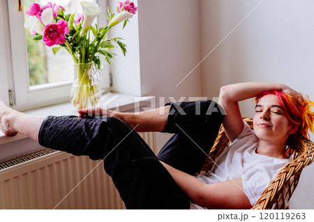 Relaxed young Caucasian woman with colorful hair sitting comfortably in wicker chair. Enjoying quiet moment by window with bouquet of vibrant flowers in vase. Tranquility and peace. 120119263