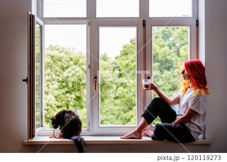 Young woman with colorful hair seated by window holds drink, cat plays nearby. Serene domestic, relaxation, leisure, and connection with yourself. Peacefulness and calm, enjoying simple moments. 120119273