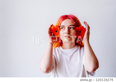 Young Caucasian woman with pink and orange hair holds vibrant gerbera flowers like headphones, expression is thoughtful and playful, radiating a sense of creativity and individuality. 120119274