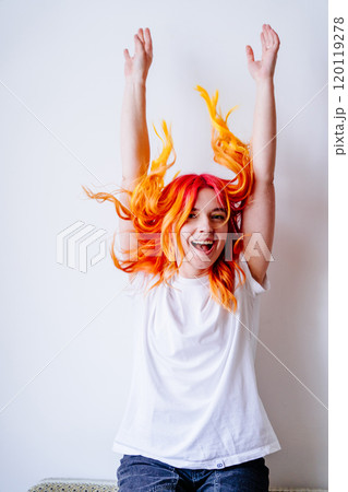 Young Caucasian woman in white t-shirt and jeans with colorful red and orange hair joyfully throwing hands in air indoors. Positive energy, playful expression and dynamic pose, white background 120119278