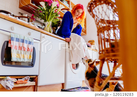 Caucasian woman wearing blue sweater with vibrant red hair sitting in cozy kitchen, interacting with dog. Bright flowers, homely interior. Domestic life, and companionship between human and pet. 120119283