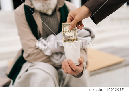 Closeup cropped shot of unrecognizable kind man giving paper money to senior homeless beggar male sitting on urban street, symbolizing hope and generosity in face of poverty and unemployment. Closeup cropped shot of unrecognizable kind man giving paper money to senior homeless beggar male sitting on urban street, symbolizing hope and generosity in face of poverty and unemployment. 120120574