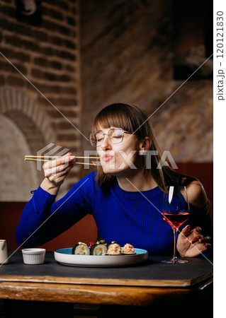 Smiling woman enjoying sushi with chopsticks, paired with a glass of rose wine in a cozy restaurant Smiling woman enjoying sushi with chopsticks, paired with a glass of rose wine in a cozy restaurant 120121830