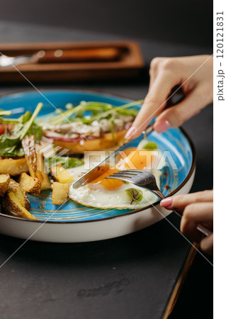 Close-up of hands cutting into a sunny-side-up egg on a breakfast plate with potatoes and greens Close-up of hands cutting into a sunny-side-up egg on a breakfast plate with potatoes and greens 120121831