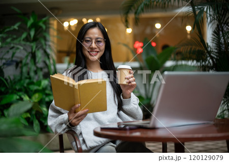 A beautiful, relaxed Asian woman in a cozy sweater enjoys coffee while reading at an outdoor table. 120129079