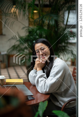 A beautiful Asian woman smiles at the camera while sitting at an outdoor table at a coffee shop. 120129116
